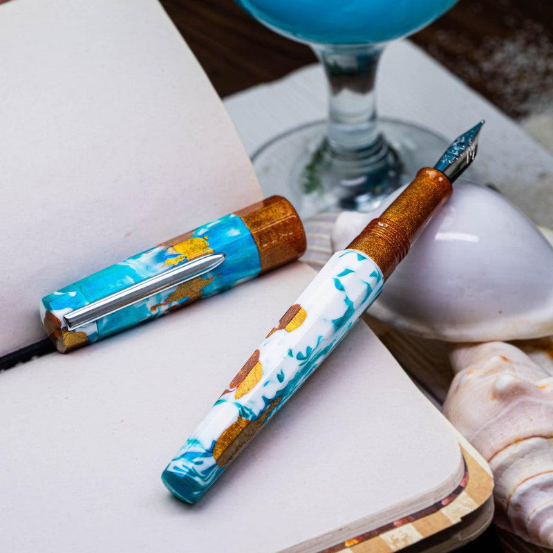 BENU Euphoria Blue Hawaiian Fountain Pen - Pen Without Cover On Top Of Notebook With A Glass Of Blue Hawaiian | EndlessPens